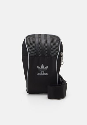 Pochette bandoulière noire Adidas avec bordure grise, trois bandes sur le dessus, logo Trèfle, fermeture éclair et sangle réglable enroulée à côté.
