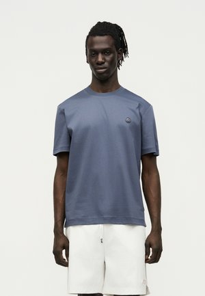 Homme à la peau foncée et aux dreadlocks portant une chemise bleue à manches courtes et un short blanc, debout devant un fond clair uni.