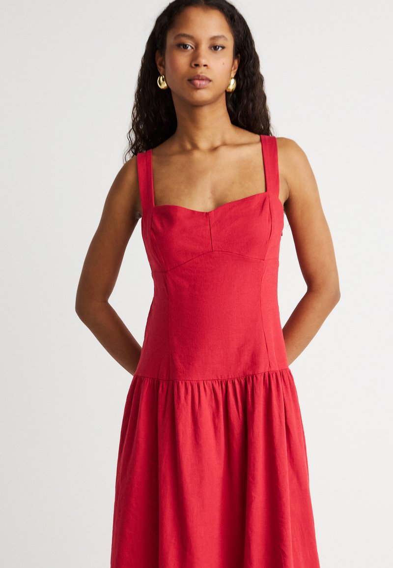 Jeune femme aux longs cheveux bouclés porte une robe rouge sans manches avec un décolleté carré et une jupe froncée, debout devant un fond clair.