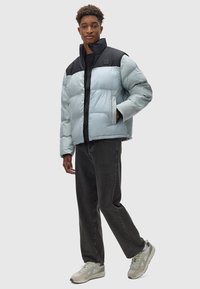 Helle blaue Steppjacke mit schwarzen Schultern, Reißverschluss vorne und Seitentaschen; kombiniert mit dunklen Jeans und hellen Turnschuhen. Sanfte Textur.