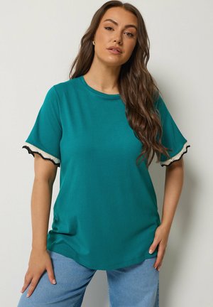 Kobieta o długich brązowych włosach, ubrana w niebieskozieloną bluzkę z krótkim rękawem z czarno-białą lamówką na rękawach oraz jasnoniebieskie dżinsy, stojąca na jednolitym tle.