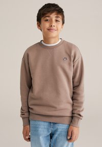 Rövid barna hajú fiú barna pulóverben, amelyen egy kis hímzett logó látható, és világoskék farmerben, egyszerű háttér előtt áll.