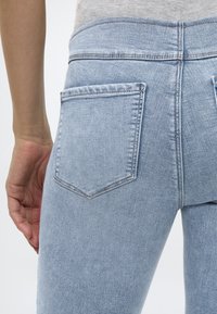 Közeli kép egy személyről, aki világoskék farmernadrágot visel, a hátsó zseb látható, és a kéz a csípő mellett pihen.