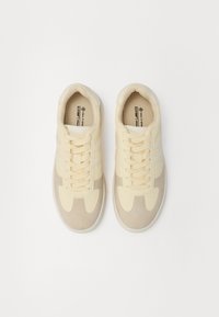Beżowe i kremowe sneakersy o teksturowanej powierzchni, z trzema białymi paskami, okrągłym noskiem i wyściełanym językiem. Związane w stylu sznurowanym.