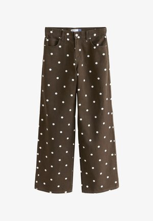 Handen die de taille van een donkere broek met witte stippen aanpassen, gecombineerd met een lichtgekleurde top die de buik en zakdetails laat zien.