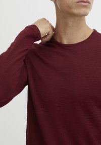 Burgunderfarget langermet skjorte med rund hals, teksturert stoff, tettsittende ermer og en glatt finish, som viser deler av skulderen og nakken.