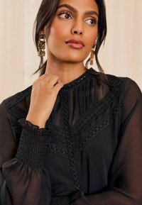 Schwarze Chiffon-Bluse mit aufwendigen Spitzenverzierungen, Falten und einem gerafften Bündchen. Goldene geometrische Ohrringe ergänzen das Outfit.