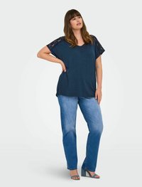 Navy top with lace short sleeves, V-neck design. Denim jeans with a straight fit and light blue wash. Model wearing heels, standing pose.