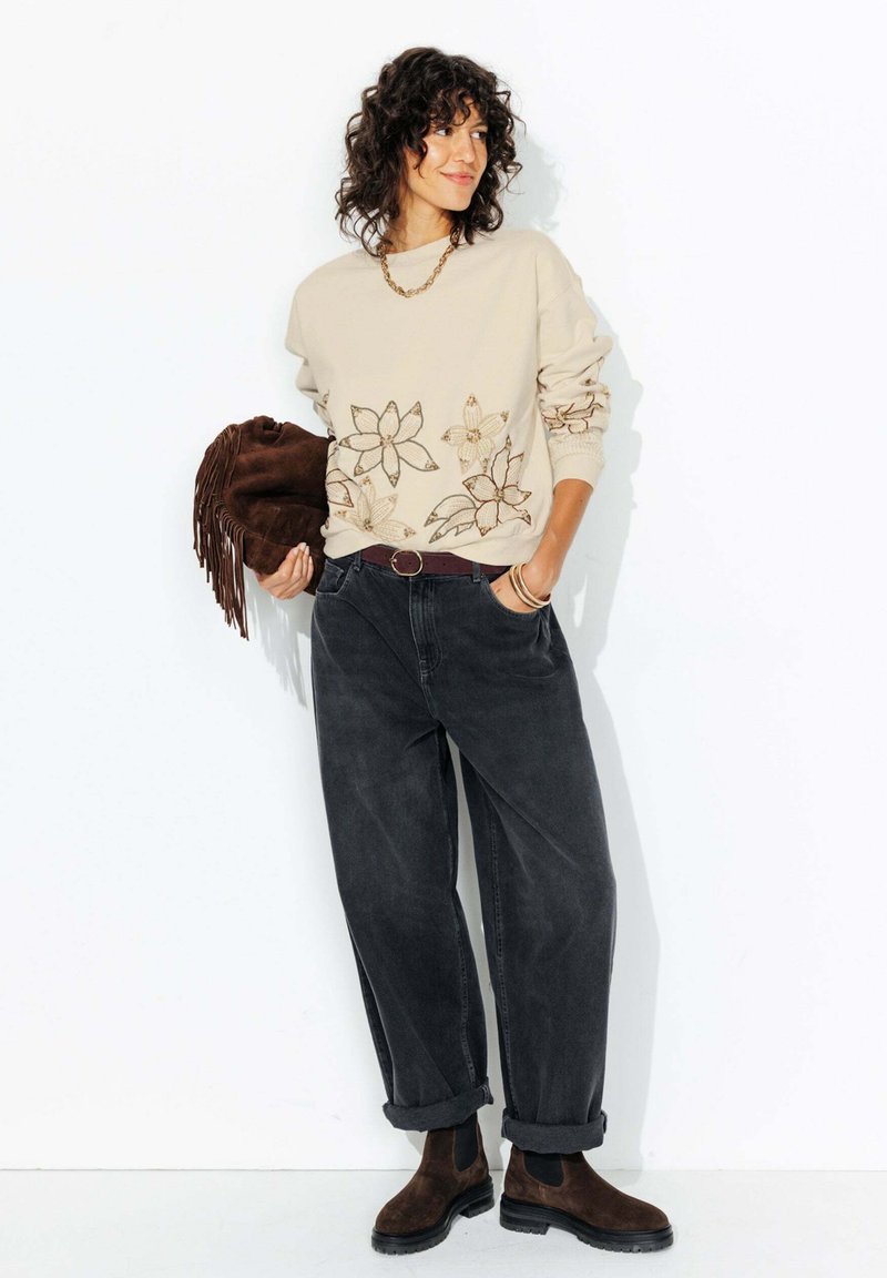 Sweatshirt beige avec broderies florales, jeans noirs ample, bottes marron et sac à franges marron, accessoirisé avec un collier en chaîne.