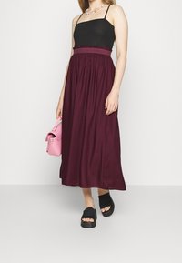 Femme portant un haut sans manches noir et une jupe bordeaux taille haute, portant un sac à main rose et des sandales à plateforme noires.