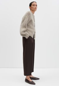 Femme portant un cardigan beige duveteux, un pantalon large marron foncé, et des ballerines pointues marron, se tenant de profil avec les mains dans les poches.