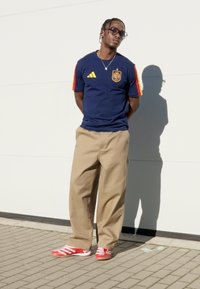 Marineblauw Adidas T-shirt met rode accenten, beige broek met wijde pijpen en rode sportschoenen met witte strepen. Bril en ketting details zichtbaar.