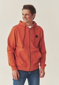 Oranje zip-up hoodie met zwarte cirkelaccent, gevoerde capuchon met gestreepte trekkoorden en twee voorzakken. Draagt blauwe spijkerbroek.