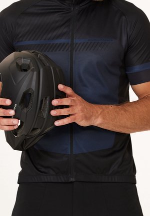 Homme en maillot de cyclisme noir et bleu marine tenant un casque de vélo noir ventilé des deux mains devant la poitrine.