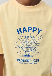 Maglietta gialla in cotone con scritta grafica blu "HAPPY PRETZEL" e immagine di un personaggio di pretzel cartone animato con le braccia.