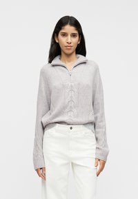 Ανοιχτό γκρι πουλόβερ με γιακά μισή ζώνη, υφασμένο με ανάγλυφο σχέδιο πλέξης και φαρδιές ριμπ μανσέτες, συνδυασμένο με λευκό παντελόνι.