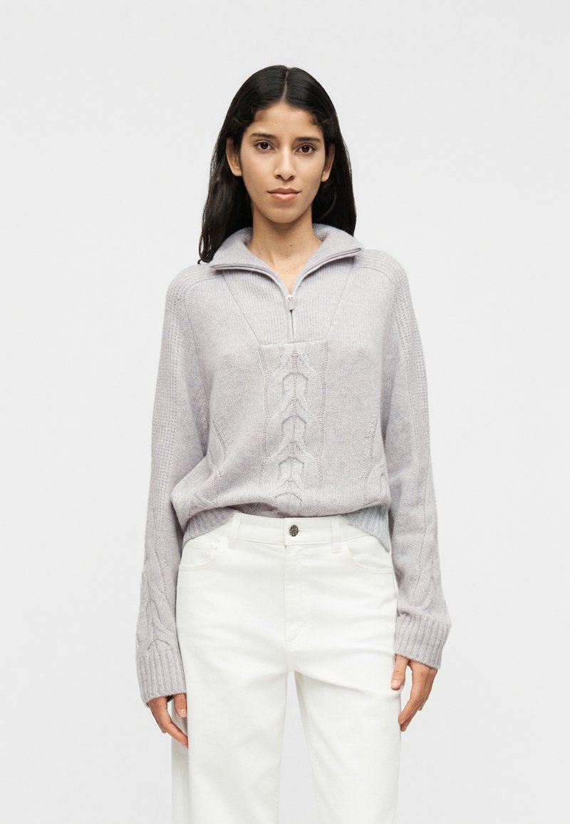 Ανοιχτό γκρι πουλόβερ με γιακά μισή ζώνη, υφασμένο με ανάγλυφο σχέδιο πλέξης και φαρδιές ριμπ μανσέτες, συνδυασμένο με λευκό παντελόνι.