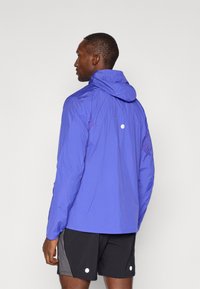 Lilla kergest vormistatud jakk kapuutsiga, millel on võrkmaterjalist detail, elastsed mansetid ja ümmargune peegelduv aktsent seljal. Mustad lühikesed püksid.