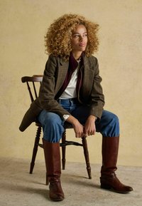 Frau mit lockigem Haar sitzt auf einem Holzstuhl und trägt ein karierter Blazer, ein weißes Hemd, eine blaue Jeans und braune knielange Stiefel vor einem beigen Hintergrund.