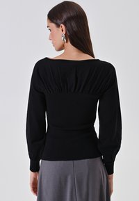 Donna con capelli scuri e lisci, che indossa una maglietta nera a maniche lunghe con dettagli arricciati sulla schiena e una gonna grigia, rivolta di spalle alla fotocamera.