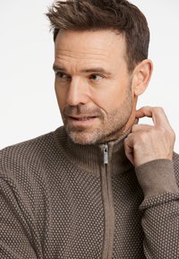 Brun zip-up tröja med ett strukturerat, prickigt mönster och ribbad krage. Tyget verkar mjukt med en åtsittande passform och dragkedja.