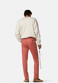 Sweatshirt beige clair, pantalons roses avec poches avant, et chaussures marron. La silhouette tient une brindille verte. Vue de dos, fond neutre.