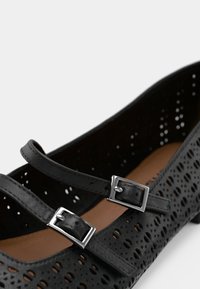 Chaussure plate noire perforée avec deux brides réglables et boucles en argent, texture mate, bout rond et semelle intérieure brune.