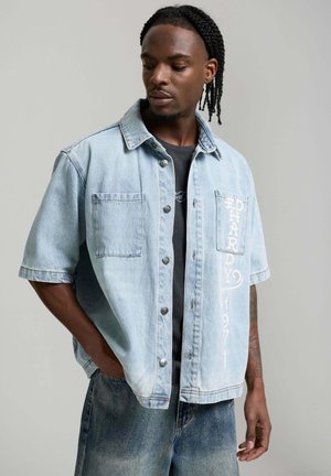 Hombre con trenzas, vistiendo una camisa vaquera de manga corta azul claro sobre una camiseta negra, y jeans azul claro, mirando hacia abajo a la izquierda.