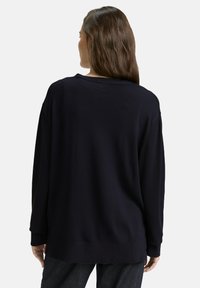 Vrouw met lang bruin haar die een losse zwarte trui en donkere broek draagt, van achteren weergegeven tegen een effen lichte achtergrond.