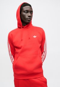 Červená mikina s kapucňou z bavlneného zmesi, s predným vreckom, bielymi rebrovanými detailmi a malým bielym logom na ľavej strane hrudníka.