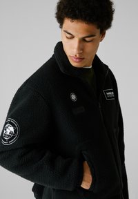 Sort fleecejakke med høj krave, lynlåslukning, forlommer og broderede patches på brystet og ærmet. Tekstureret stof.