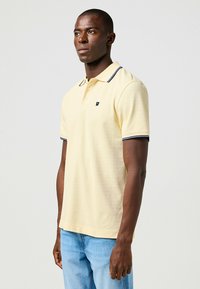 Polohemd aus leichtem gelben Baumwollstoff mit navyblauem geripptem Kragen und Ärmeln, mit einem kleinen Logo auf der linken Brust.