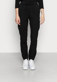 Mustat jogger-housut, joissa on nyöri vyötäröllä, sivutaskut, joustavat hihnat, yhdistettynä valkoisiin tennareihin. Sujuva rakenne ja rento muotoilu.