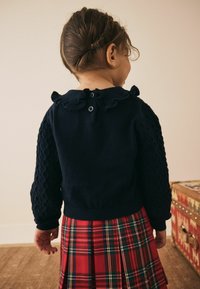 Mørkeblå strikket trøje med strukturerede ærmer og flæsekrave, med knaplukning bagpå; parret med en rødternet nederdel.