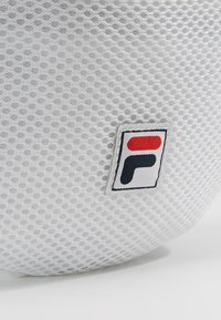 Tissu en maille texturée blanche avec un petit patch logo rectangulaire arborant une lettre stylisée rouge et noire sur fond blanc.