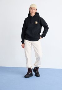 Black hoodie with a front pocket and gold logo, paired with light striped pants and black lace-up boots with red accents, standing on blue floor.