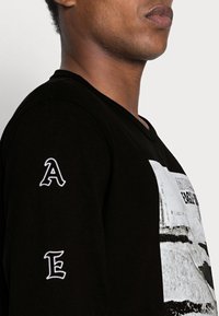 Musta pitkähihaine paita, jossa on valkoinen graafinen printti etupuolella, joka esittää suurta tekstimuotoilua. Koristeina valkoiset kirjaimet 'A' ja 'E' hihoissa.