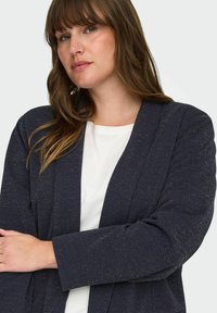 Námořnický blazer s jemnými zlatými flíčky, dlouhými rukávy a otevřeným designem vpředu, pod ním bílé tričko. Hladká textura.