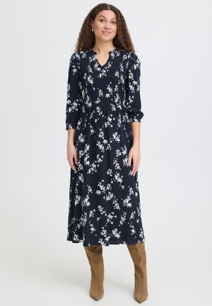 Femme aux cheveux bouclés portant une robe midi noire à fleurs et des bottes montantes en daim beige, debout devant un fond uni.