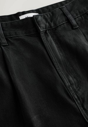 Nahaufnahme des Taillenbunds von schwarzen Jeans mit Gürtelschlaufen, sichtbarer Reißverschlussnaht und teilweise sichtbarem weißen Etikett.