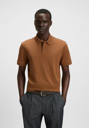Camisa polo marrón de manga corta, con textura suave, tapeta de dos botones y logo bordado; combinada con pantalones grises ajustados y un cinturón marrón.