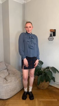 Ανοιχτό μπλε μπλούζα με βολάν μπροστά, συνδυασμένη με φούστα καρό. Μαύρα χοντρά παπούτσια και λευκές κάλτσες. Εσωτερικός χώρος με φυτά.