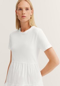 Weißes kurzes Kleid mit kurzen Ärmeln und Rundhalsausschnitt, mit einem taillierten Oberteil und einem fließenden, gestuften unteren Teil. Weicher, leichter Stoff.