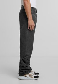 Muž v volných černých džínách a bílých, béžových a černých teniskách stojící v profilu proti jednoduchému šedému pozadí.