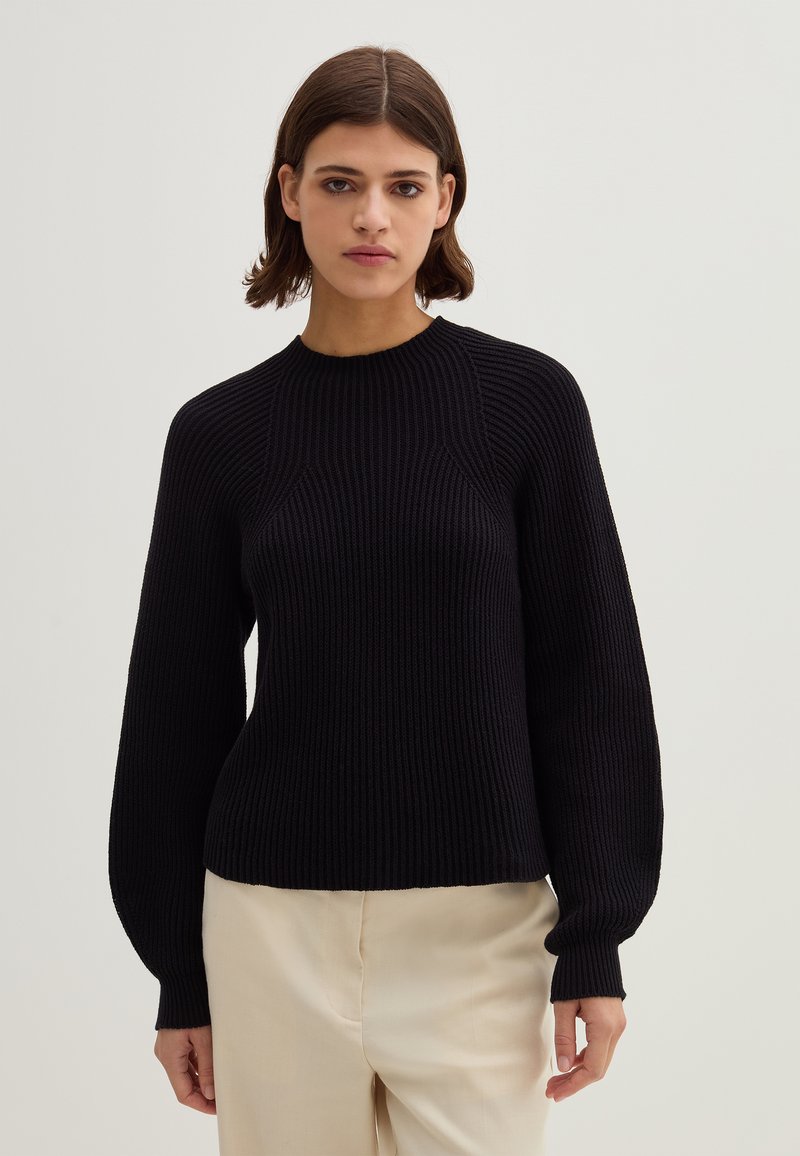 Frau mit kurzem braunem Haar, die einen schwarzen, gerippten Langarmpullover und eine hellbeige Hose trägt und vor einem neutralen Hintergrund steht.