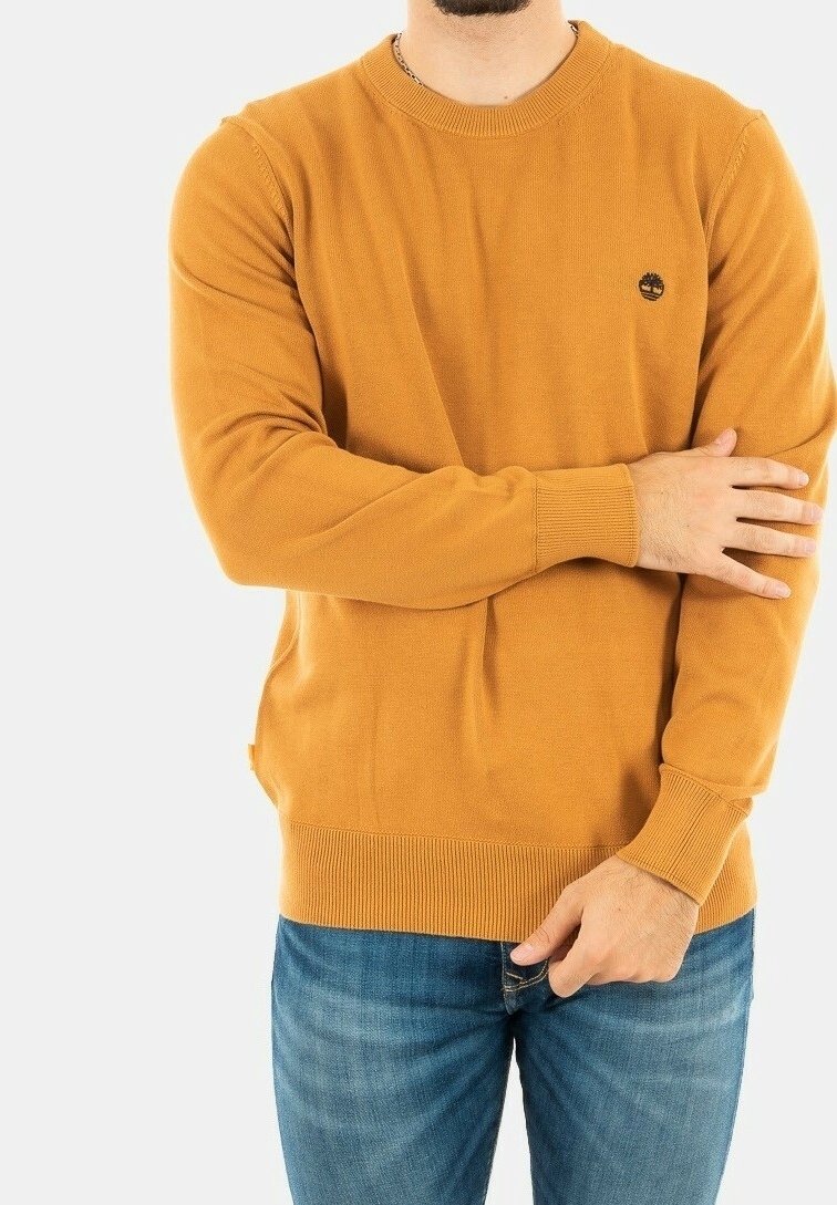 Pull en tricot jaune moutarde avec un col rond, des poignets et un ourlet côtelés, arborant un petit logo noir sur la poitrine. Porté avec un jean bleu.