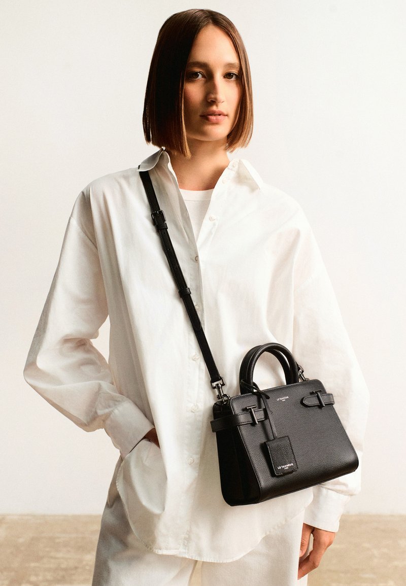 Sac en cuir noir avec double anses supérieures, une bandoulière détachable et une finition texturée. Dispose d'une quincaillerie minimaliste et d'une petite étiquette à l'avant.