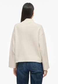 Kremfarget strikkegenser med høy krage, vide ermer og ribbet kant. Kombinert med mørkeblå denimjeans.