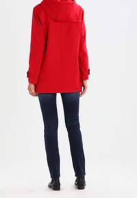 Rode capuchonjasje met een rechte snit, zichtbare naden en afgewerkte manchetten met knopen. Draagt over donkerblauwe spijkerbroeken met zwarte enkellaarzen.