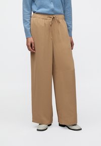 Μπεζ παντελόνι φαρδιάς γραμμής με ελαστική μέση και ρυθμιζόμενο κορδόνι, κατασκευασμένο από απαλό ύφασμα. Φορεμένο με γαλάζια ζακέτα και παπούτσια.